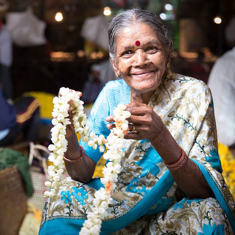 An elderly market woman smiles and shows off a necklace of flowers.