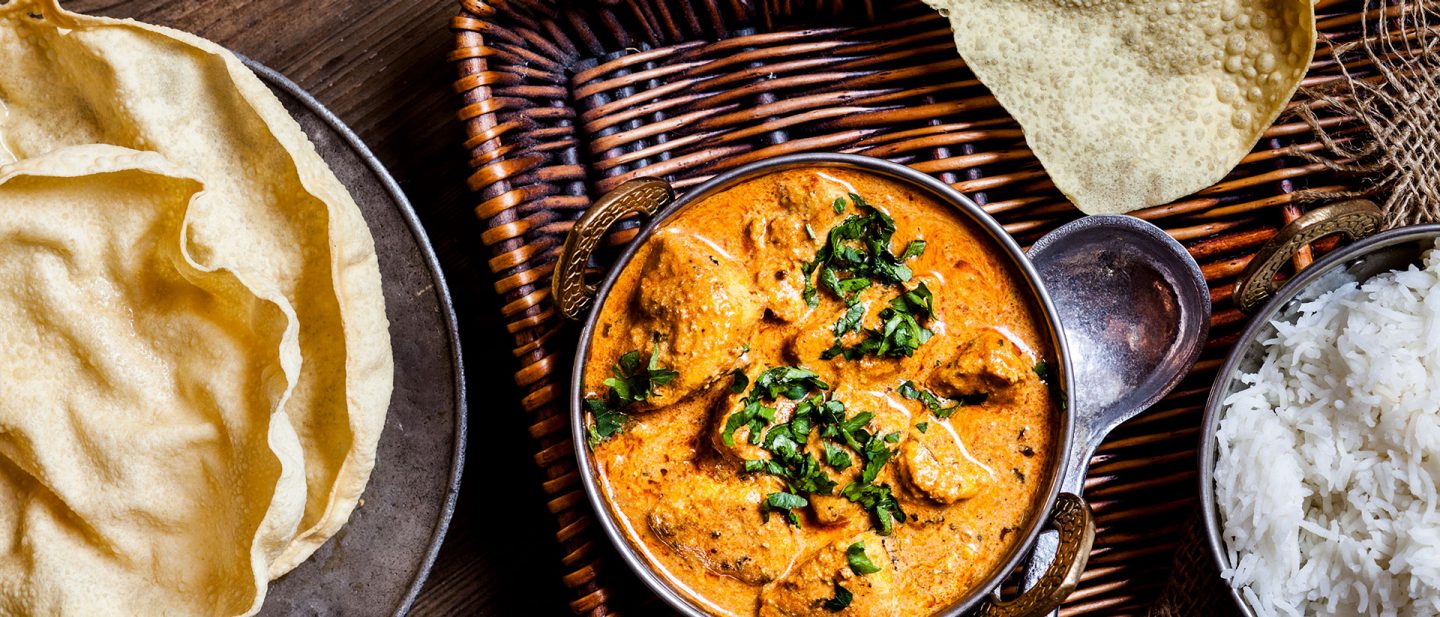 Picture of a bowl of butter chicken on a basket tray, with rice and thin flatbread next to it