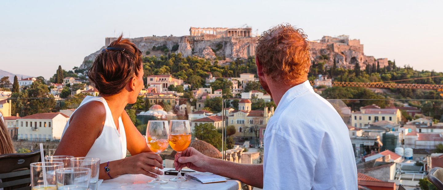 Two people sit at a table, each with a cocktail in their hand, looking at the Acropolis in the distance.
