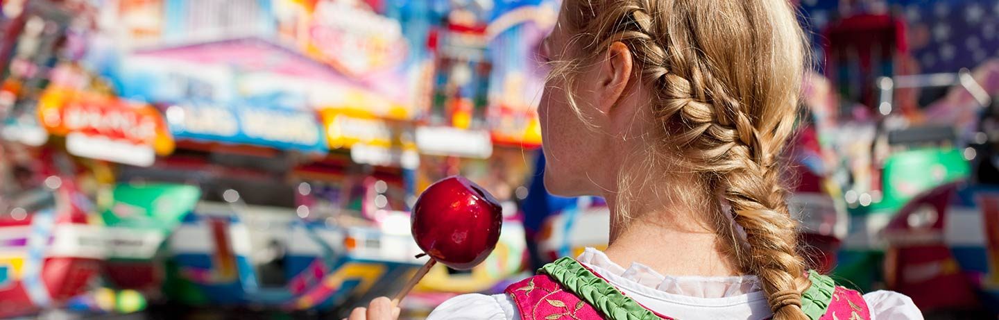 Image of a girl with braided hairstyle and dirndl from behind, eating a candied apple, in the background colorful rides. 
