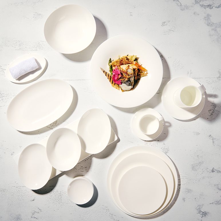 Plain white plates, cups and saucers in various sizes and shapes on a white background. A high-quality main course is arranged on one of the plates.