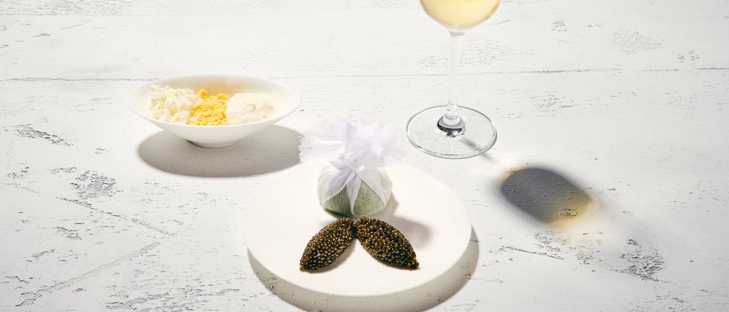 In the foreground: caviar on a white plate with lemon, packed in a sachet, background: a bowl with garnish and a champagne glass, reflected on the table.