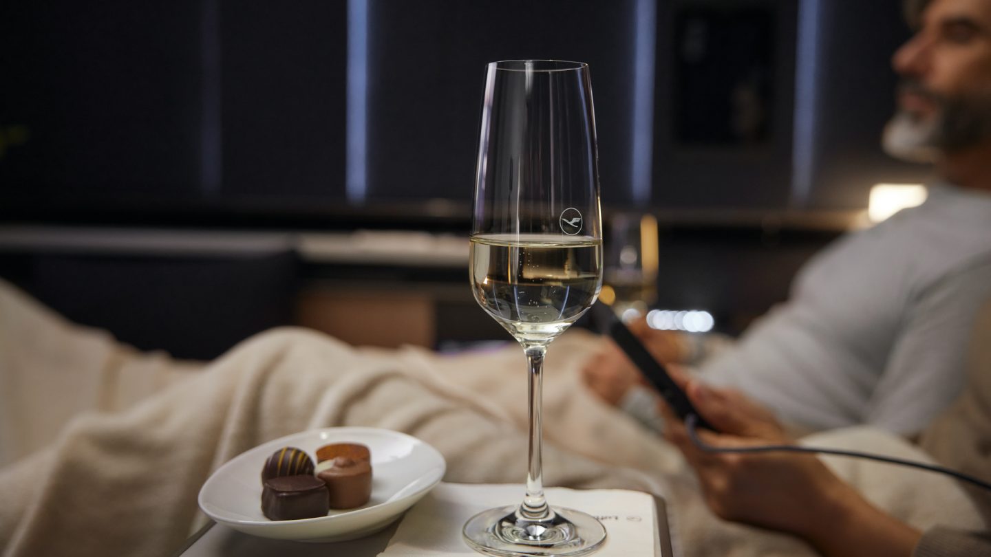 In the background is a man with a beard lying in an Allegris First Class suite; in the foreground is a glass of champagne and a bowl of chocolates.