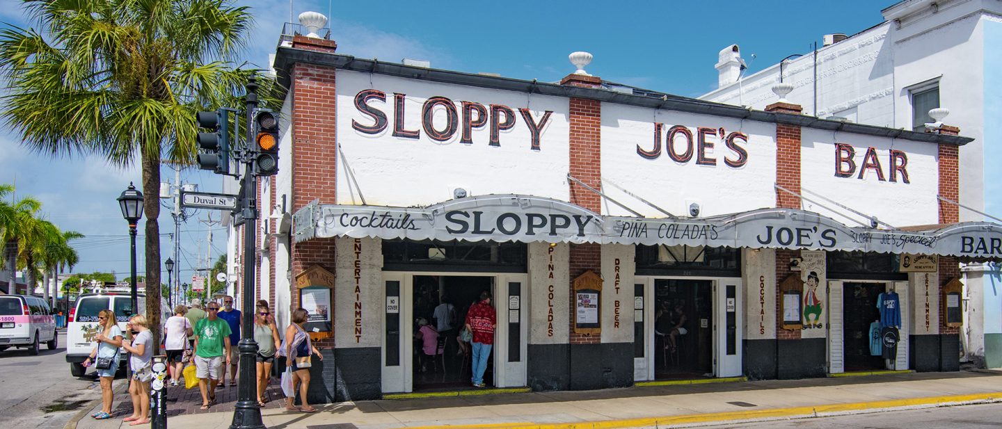 Façade of Sloppy Joe's Bar in Key West, next to it people walk along the street past a palm tree
