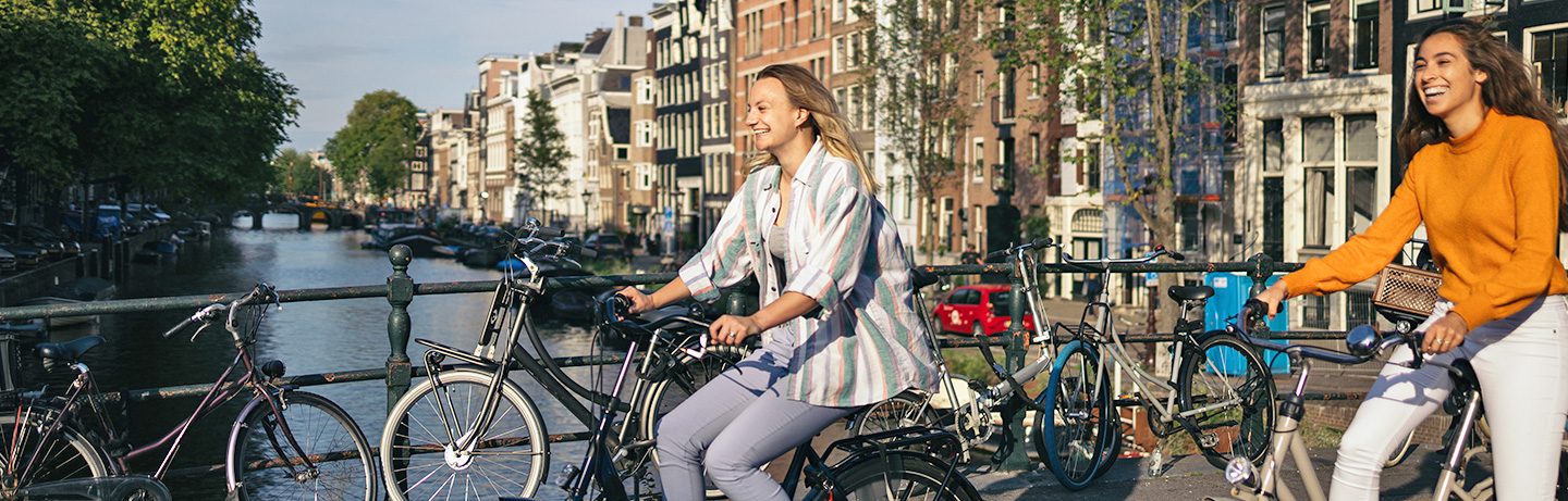 Two young women laughing as they ride their bicycles over a bridge in Amsterdam, with canals in the background