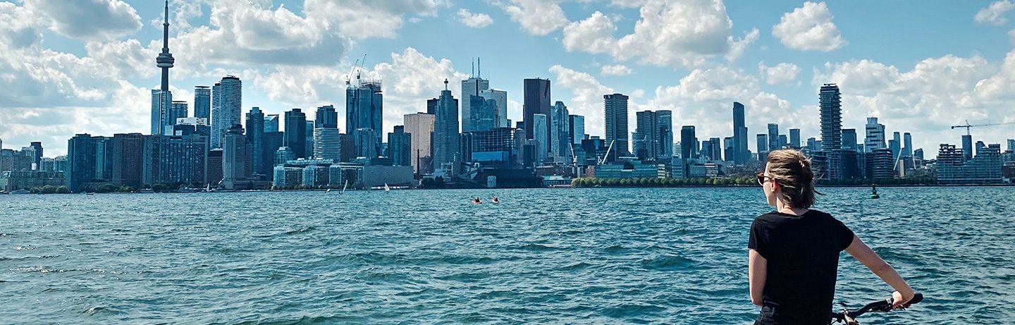 Woman with short hand stands with her back to the picture, in the background: water and the skyline of Toronto