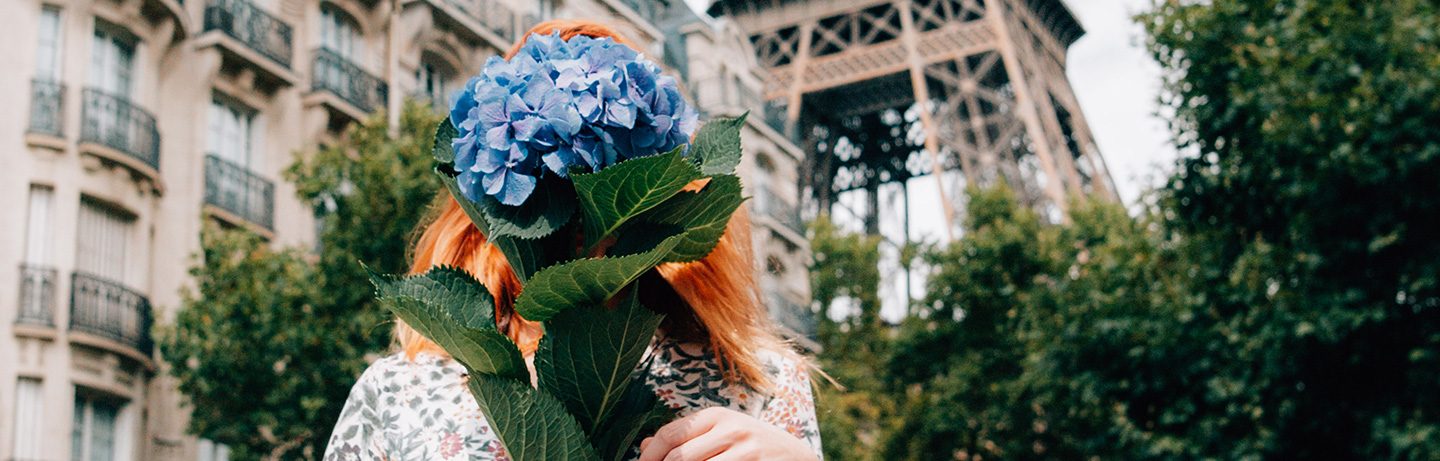 Woman with red hair hides in front of a blue hydrangea, outlines of the Eiffel Tower can be seen in the background