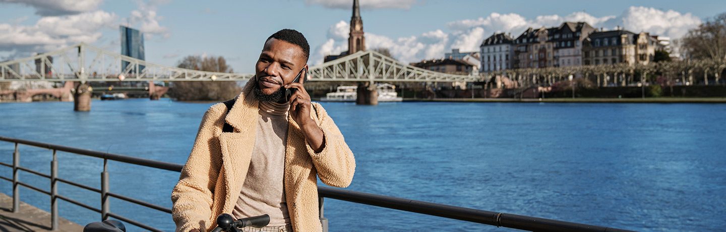 Picture of a young black man standing on the banks of the Main and talking on the phone.