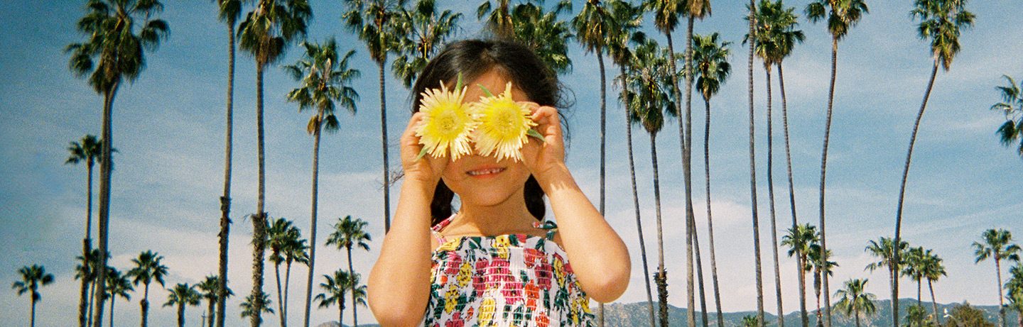 Child holds two flowers in front of their eyes, surrounded by palm trees under a blue sky