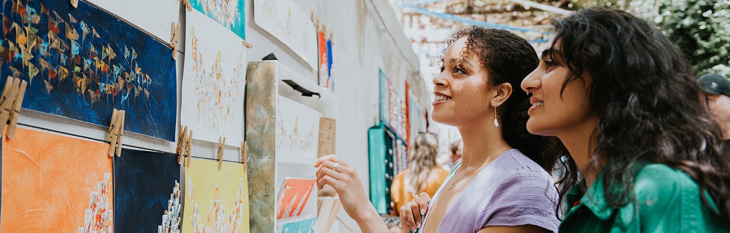 Two women smiling as they admire artworks at an outdoor exhibition
