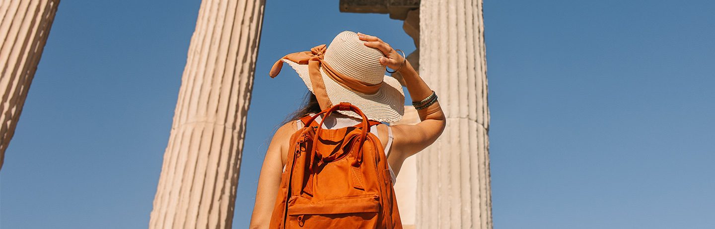 Woman with a backpack and hat looking at ancient columns under a clear blue sky