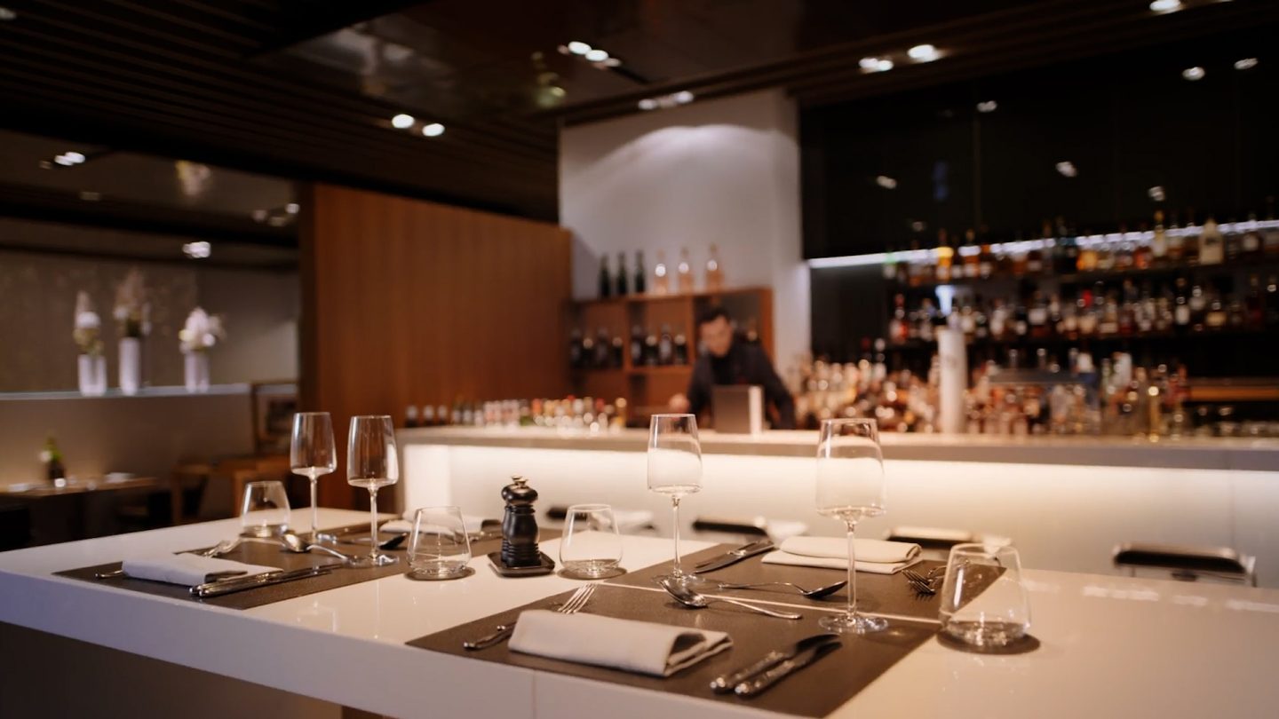 Bartender in a suit pouring red wine at a well-stocked bar.