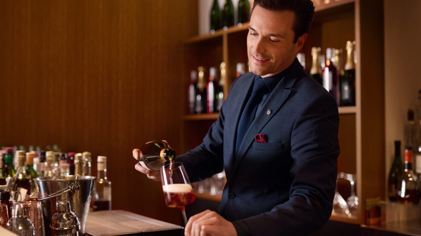 Bartender in a suit pouring red wine at a well-stocked bar.