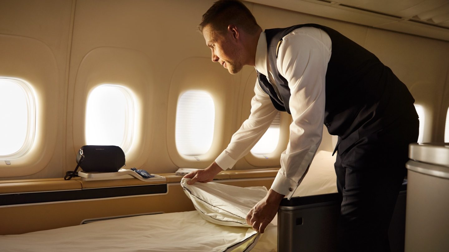 A flight attendant in uniform makes a bed in an airplane's first-class cabin.