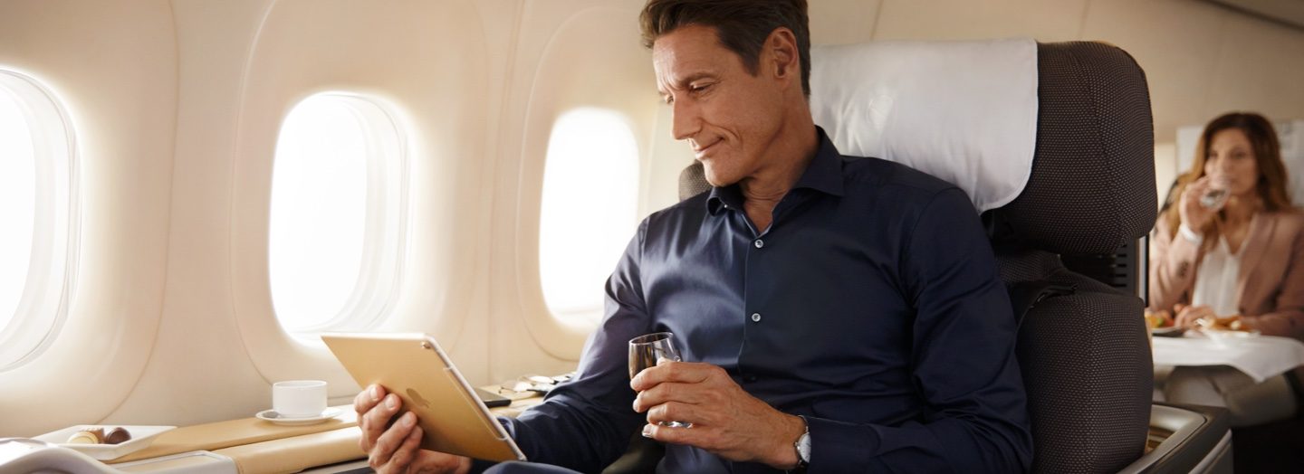 Man in business class airplane seat, holding a tablet and a drink, smiles contentedly.