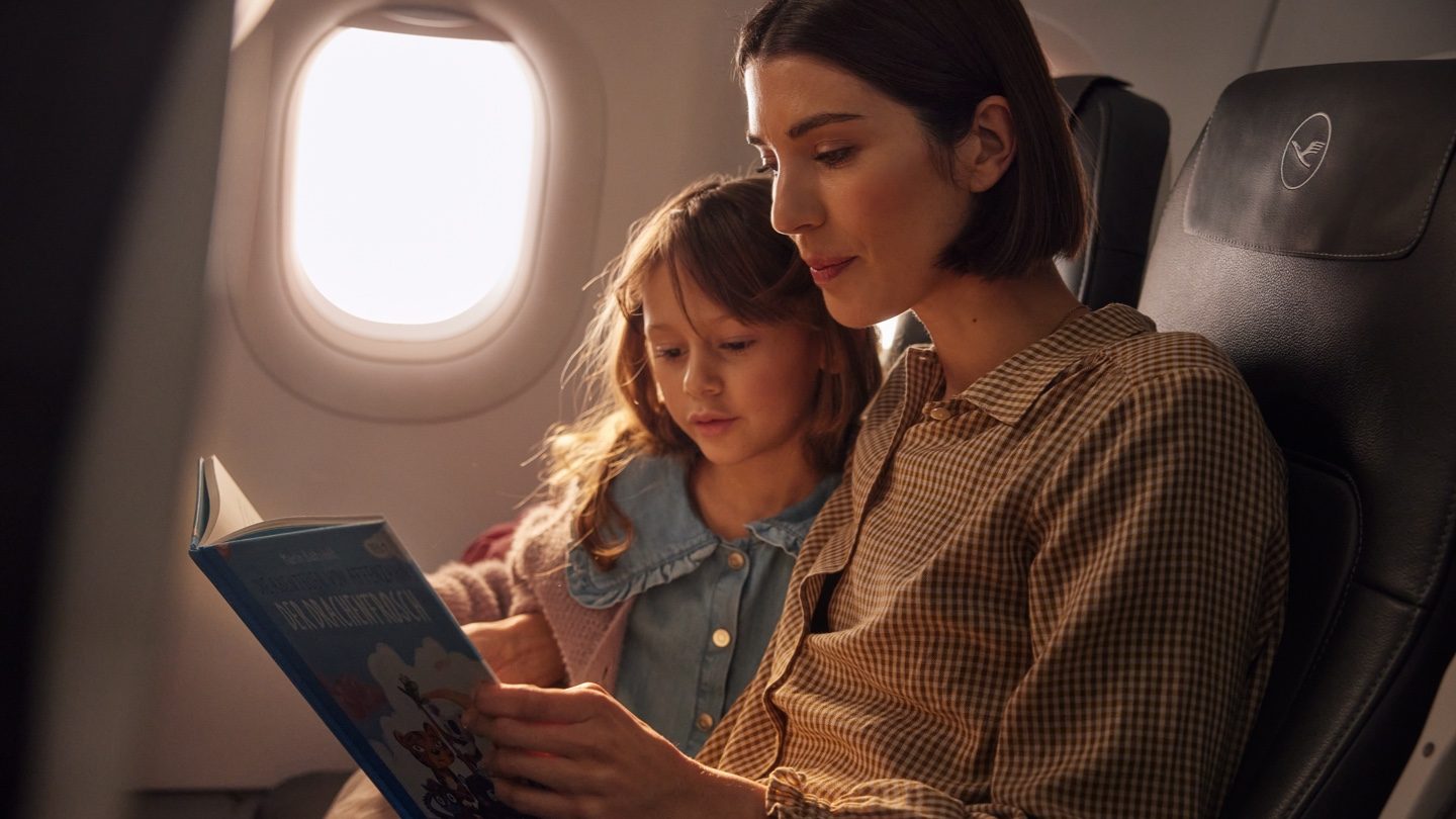 A mother and daughter are seated on an airplane, reading a book together.