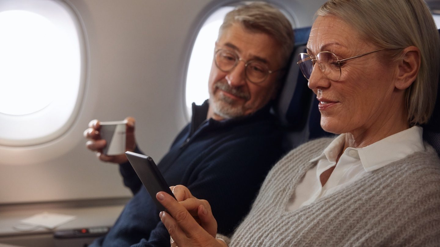 Older couple in airplane seats: woman uses smartphone, man smiles with coffee.