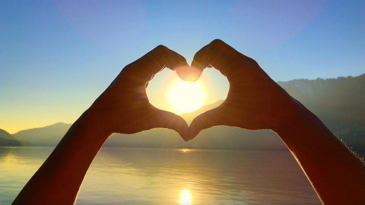 Hands form a heart shape in front of a mountain and lake landscape, with the setting sun shining through the center.