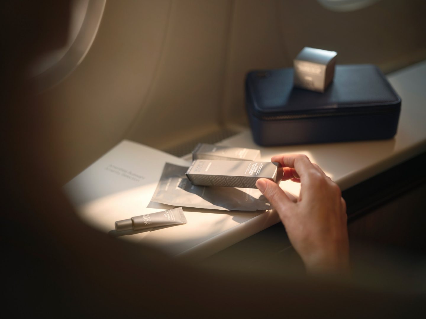 A hand arranges luxury skincare products on an aircraft's window seat tray.