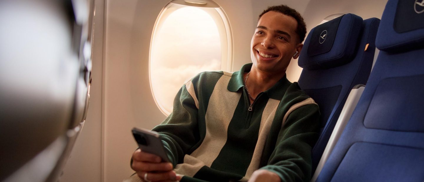 Passenger smiles, holding phone, wearing earbuds by window on a Lufthansa flight.