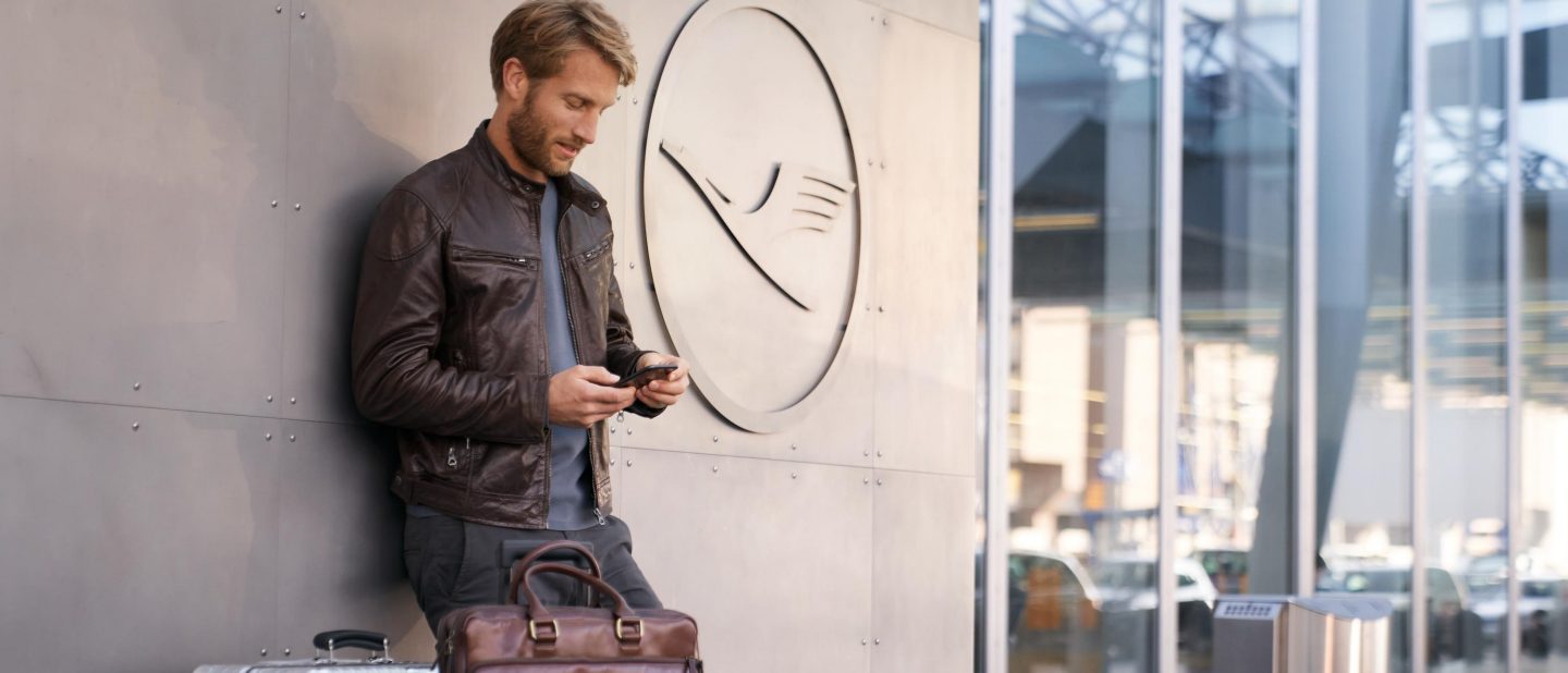 Man in a leather jacket with a suitcase standing near a check-in options sign.