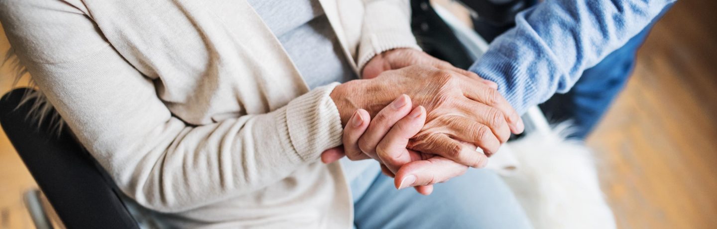 An old man and a woman in wheelchair at home, holding hands.