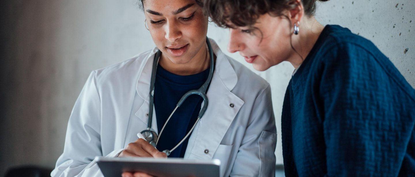A woman doctor with a stethoscope and a female patient look at a tablet together.