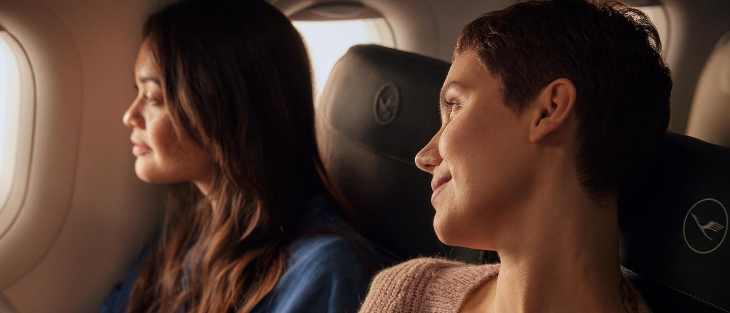 Two women relaxing on a flight. One gazes out the airplane window, the other smiles contentedly.