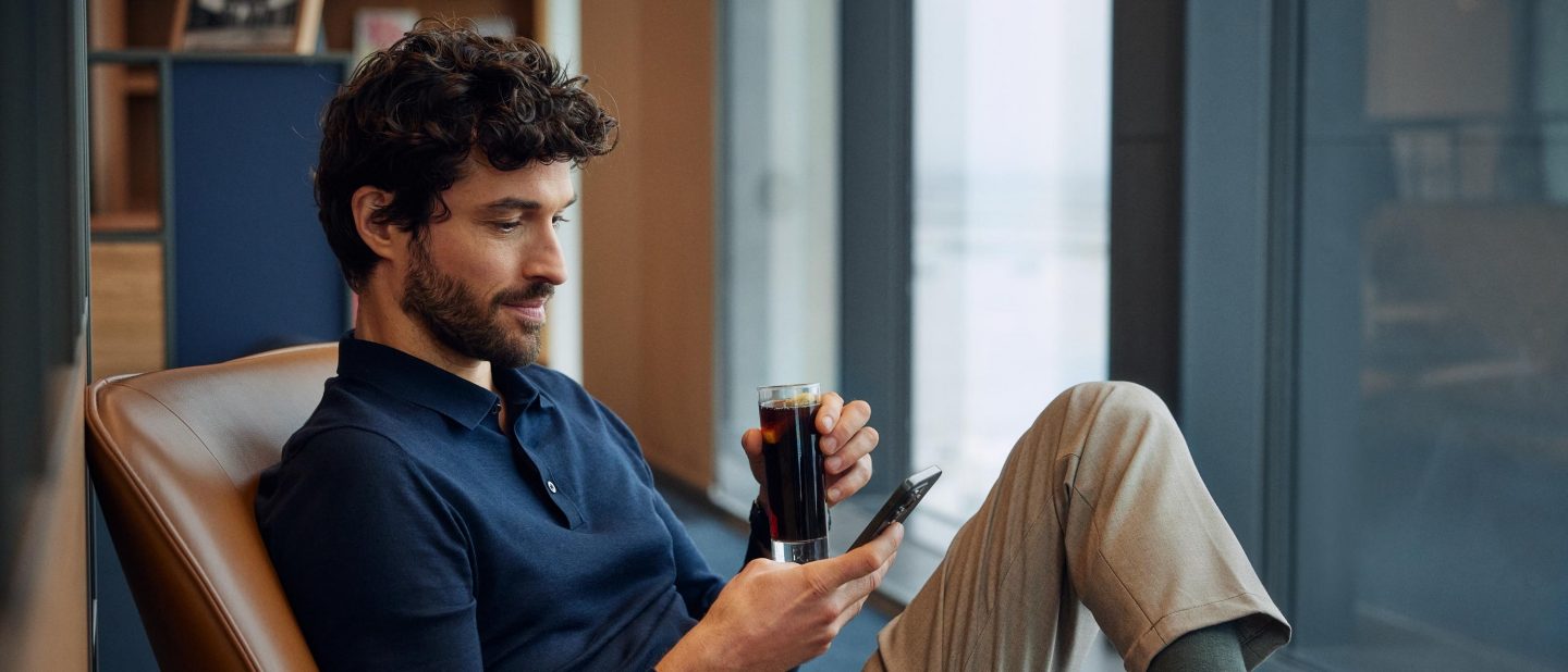 Ein Mann sitzt in der Lounge, trinkt ein Getr&auml;nk und liest etwas auf dem Handy. // A man is sitting in the lounge, sipping a drink and reading something on his cell phone.