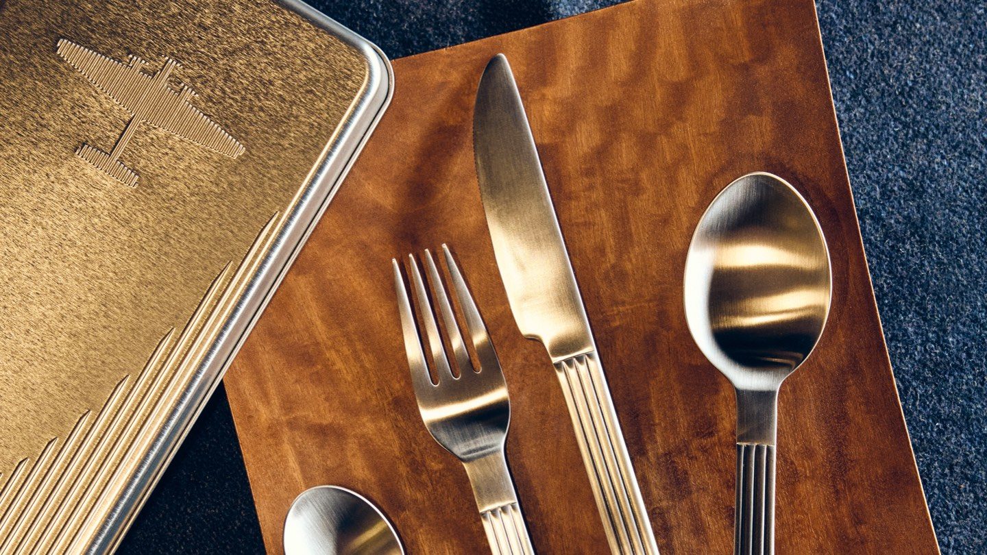 Premium airline silverware next to an embossed vintage airplane tin on a wood table.