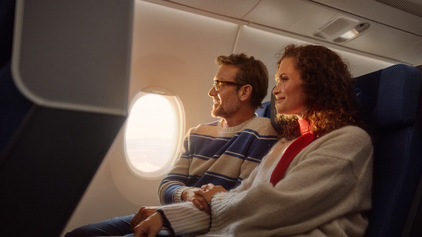 A couple holding hands and smiling while looking out an airplane window.