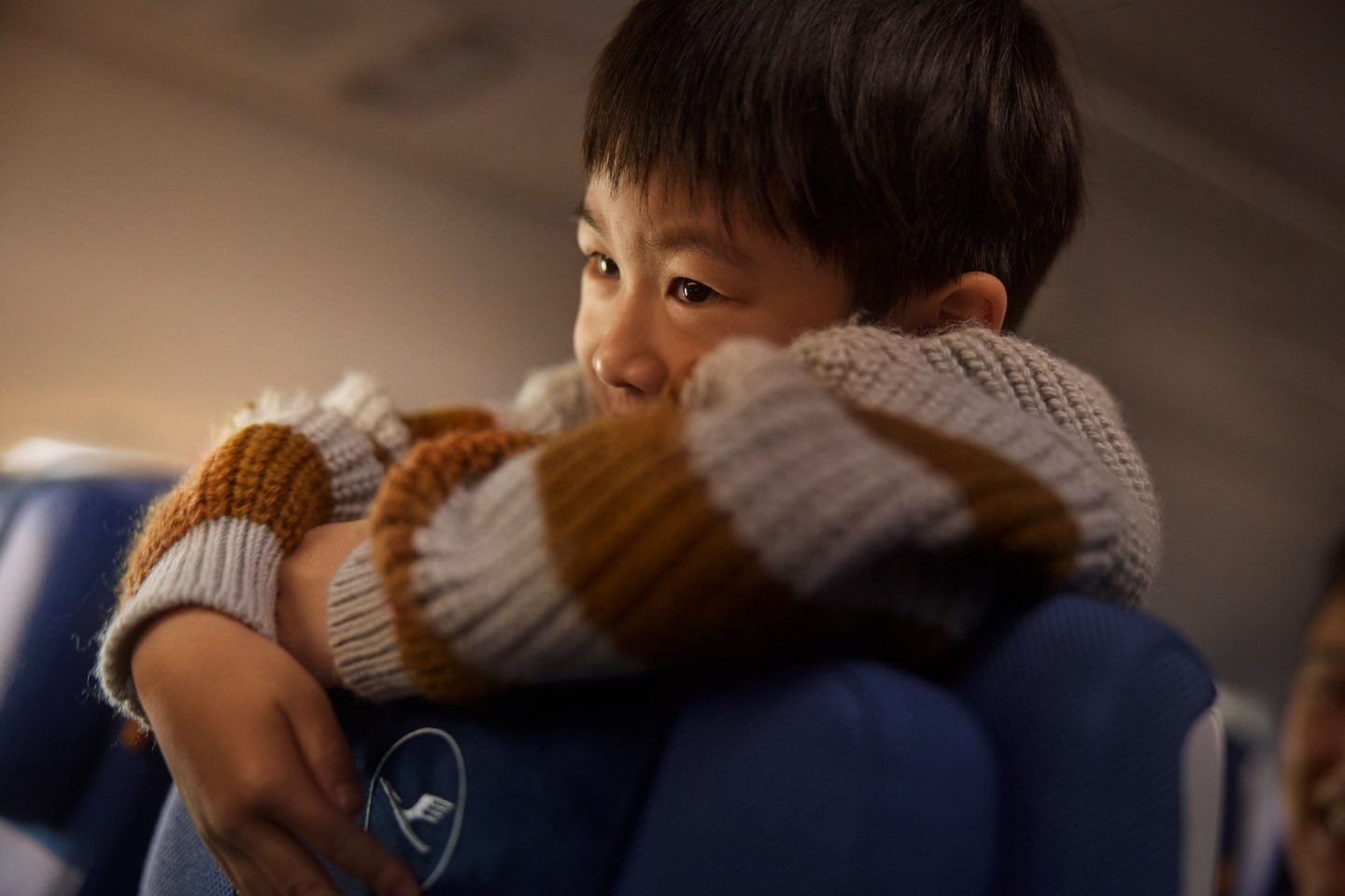 A young boy in a striped sweater smiling while resting his head on an airplane seat.