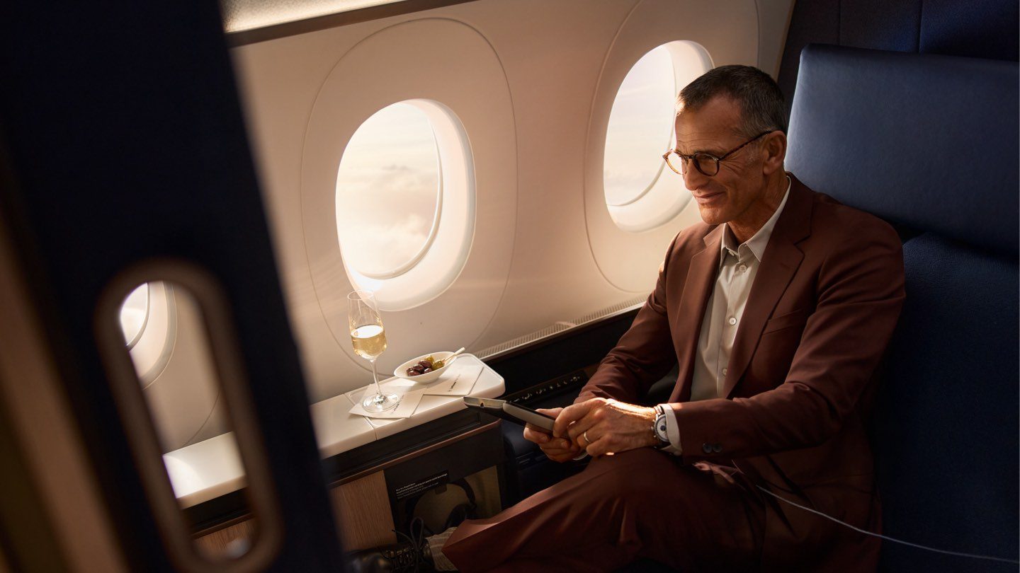 Smiling man in premium airline seat enjoys champagne, tablet, and window view.