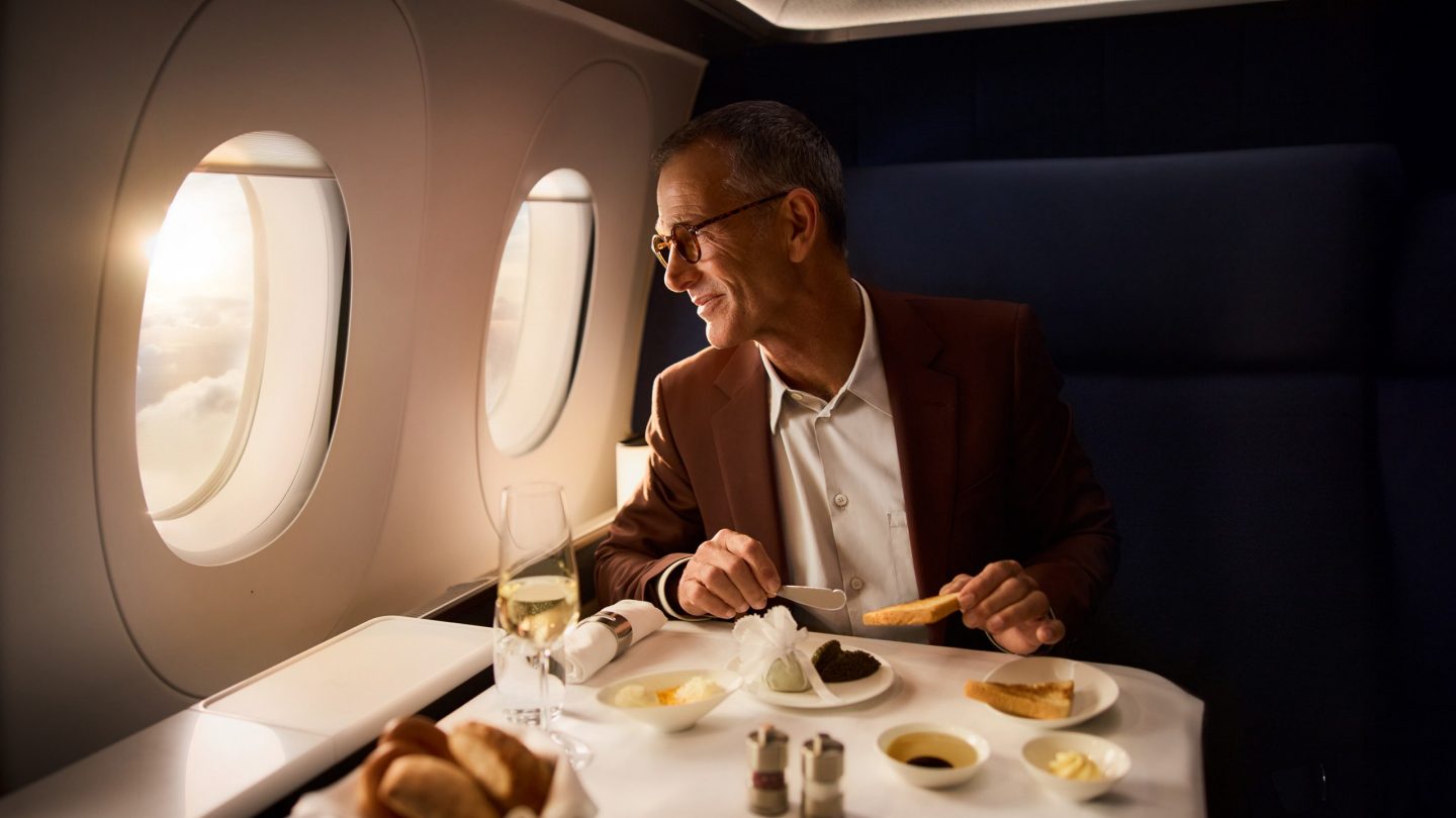 Passenger enjoying a premium meal & champagne, looking out plane window at sunny clouds.