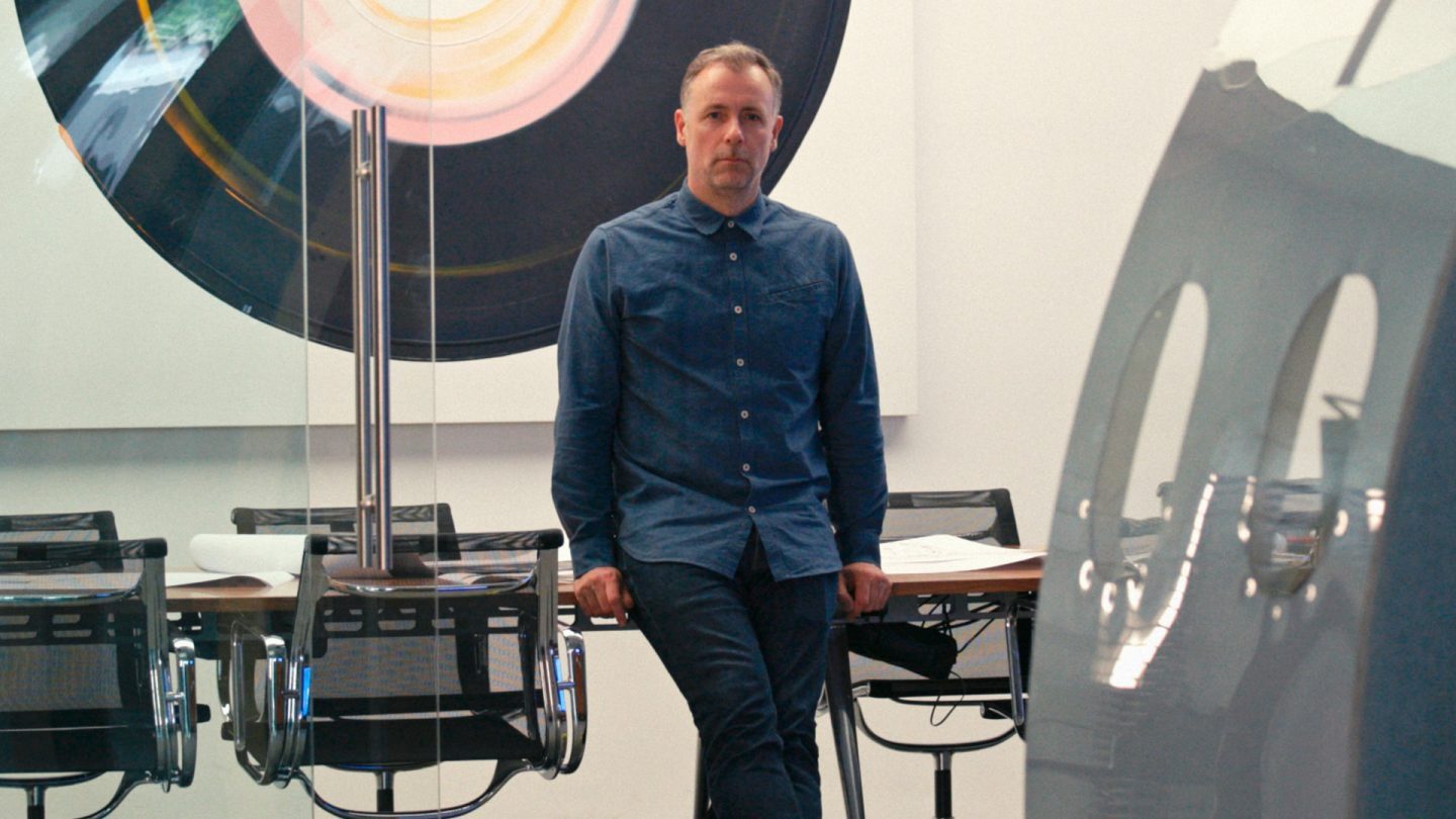 Man in blue shirt stands in an office with an aircraft structural frame.