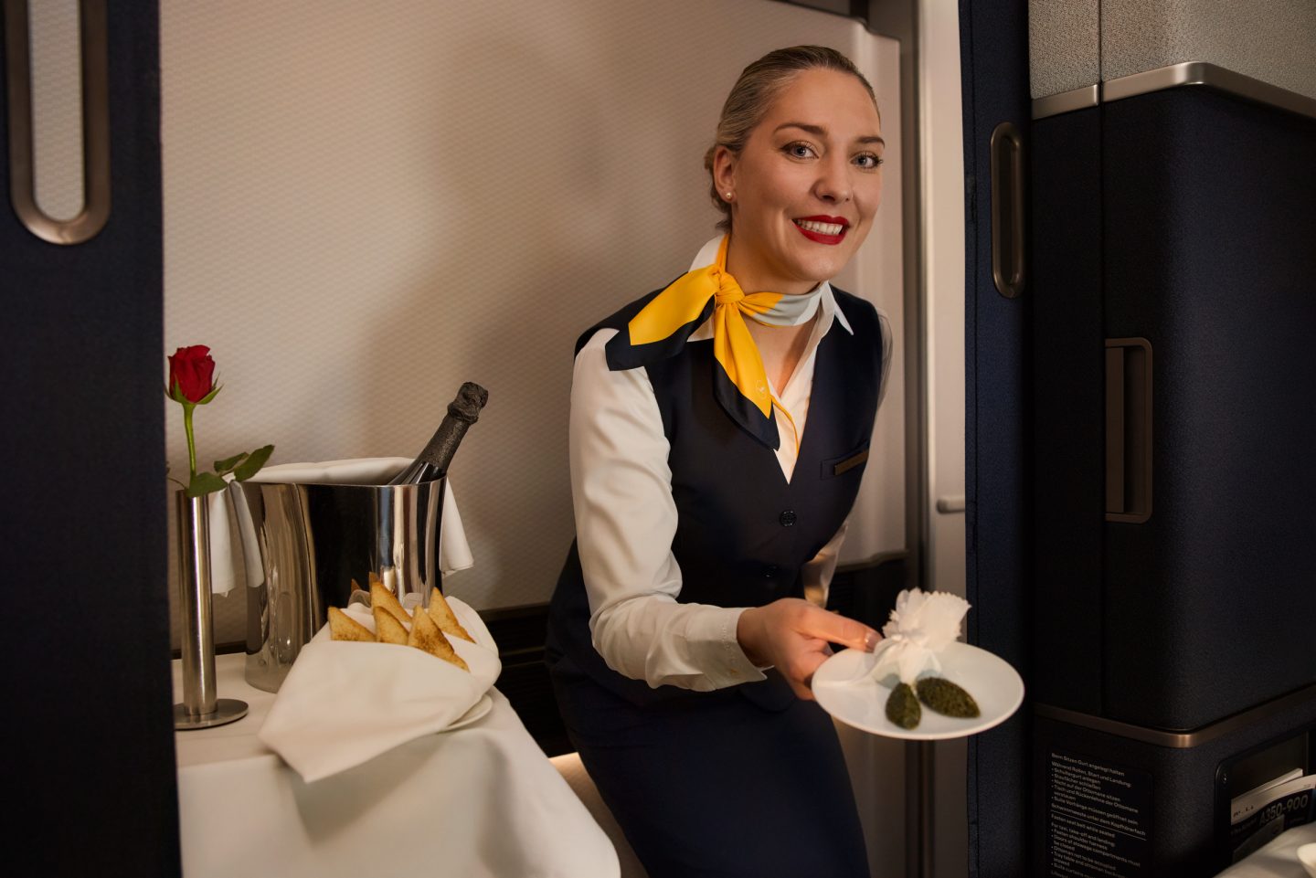 Smiling flight attendant serves a meal with champagne and roses in a premium cabin.