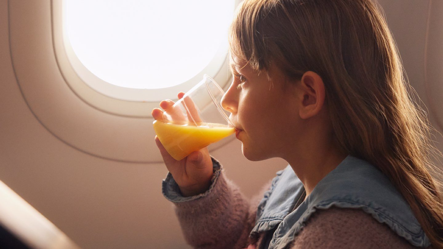 A young girl by an airplane window enjoys orange juice while reading a book.