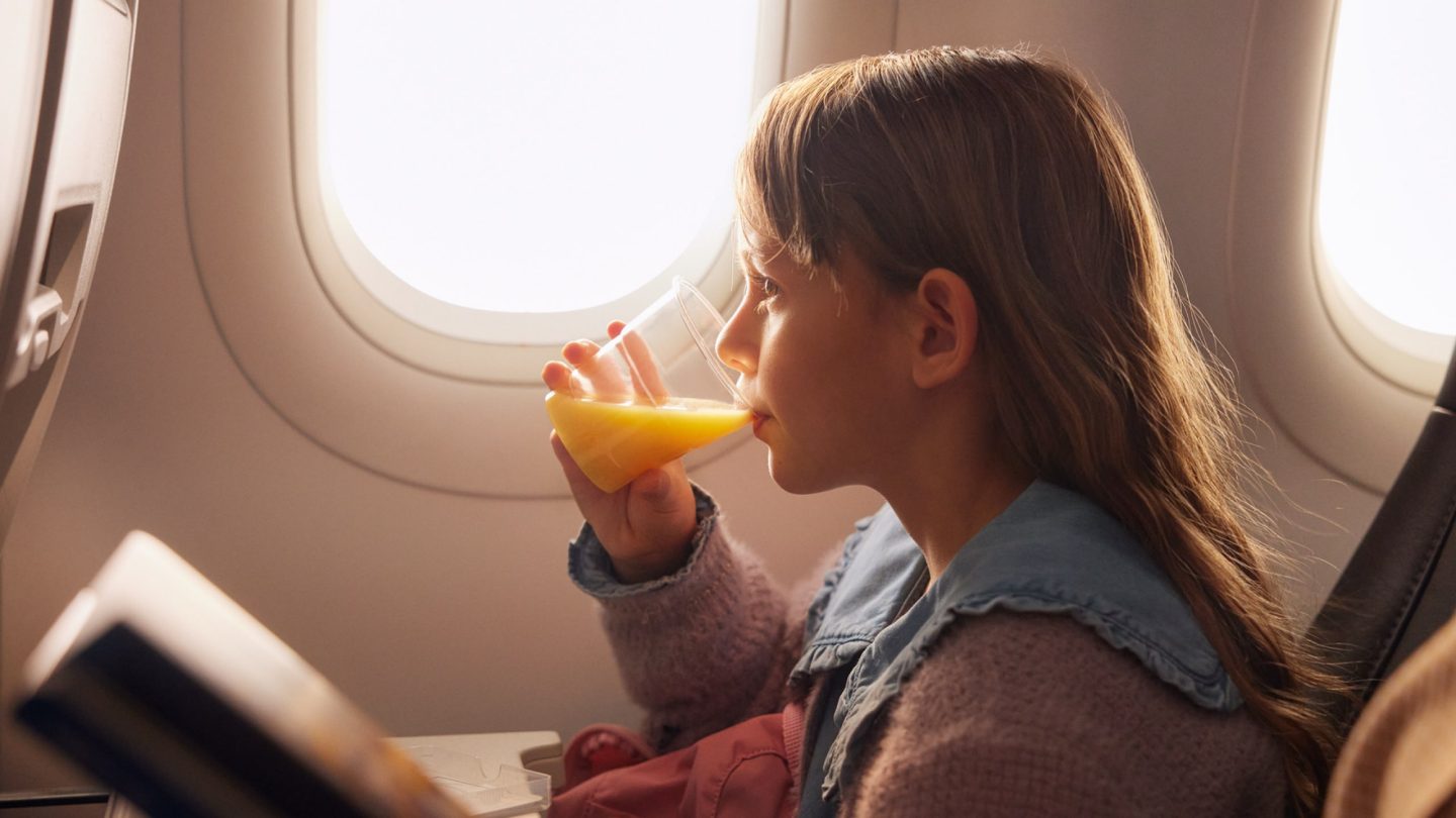 A young girl by an airplane window enjoys orange juice while reading a book.