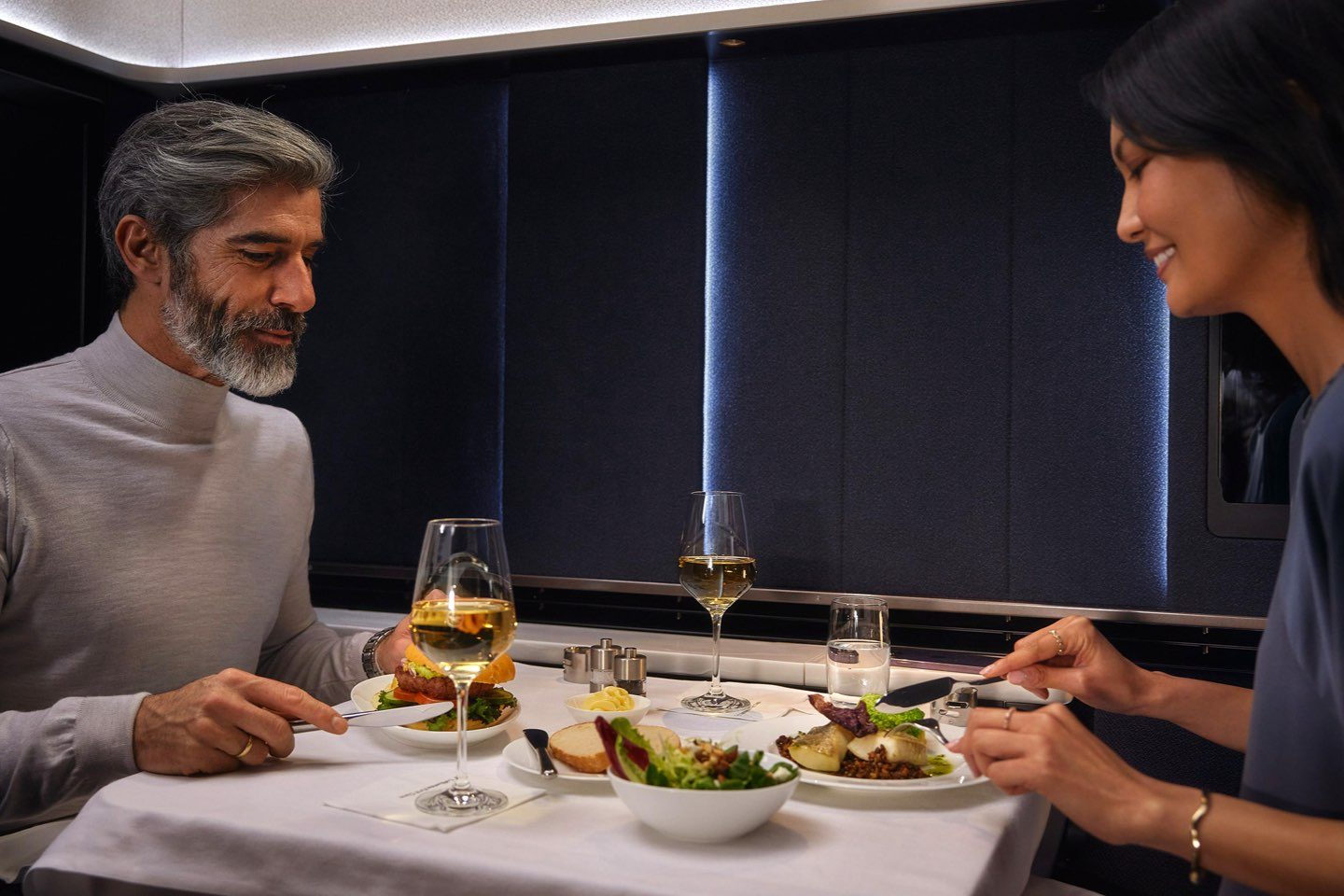 A man and woman savoring dinner in First Class while flying on an airplane.