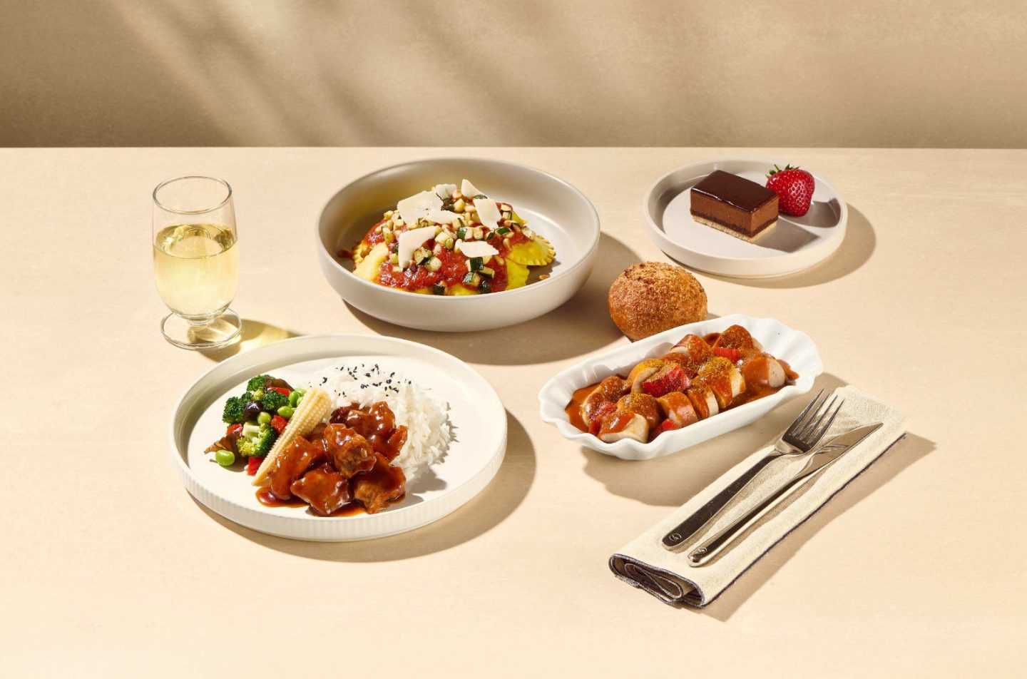 Assorted meal dishes including ravioli with tomato sauce, rice with vegetables and meat, sliced sausage with sauce, a bread roll, chocolate dessert with a strawberry, and a glass of white wine.