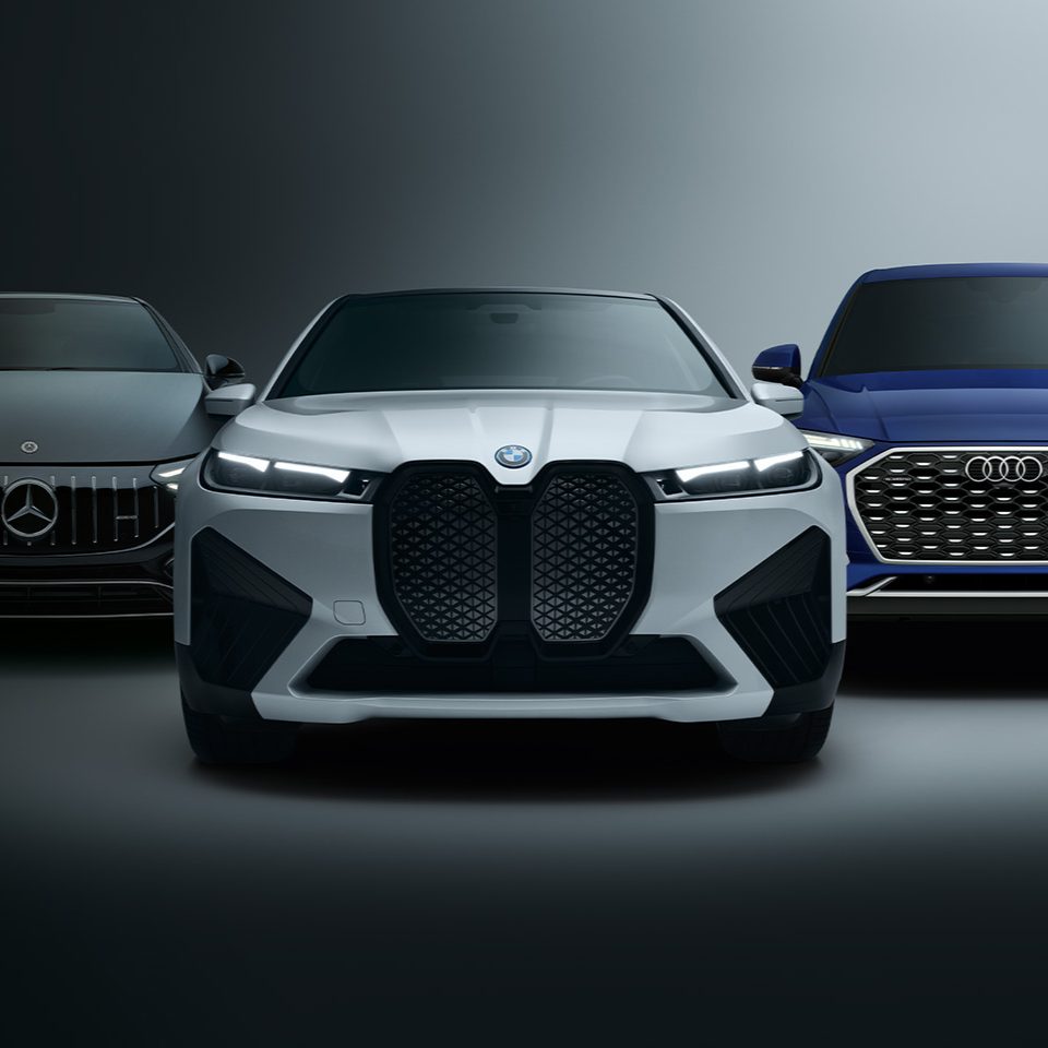 Front view of three cars from different brands against a dark background: a black Mercedes on the left, a white BMW in the center, and a blue Audi on the right.