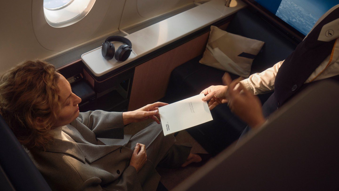 A woman in a business class seat on an airplane is handed a document by a flight attendant.