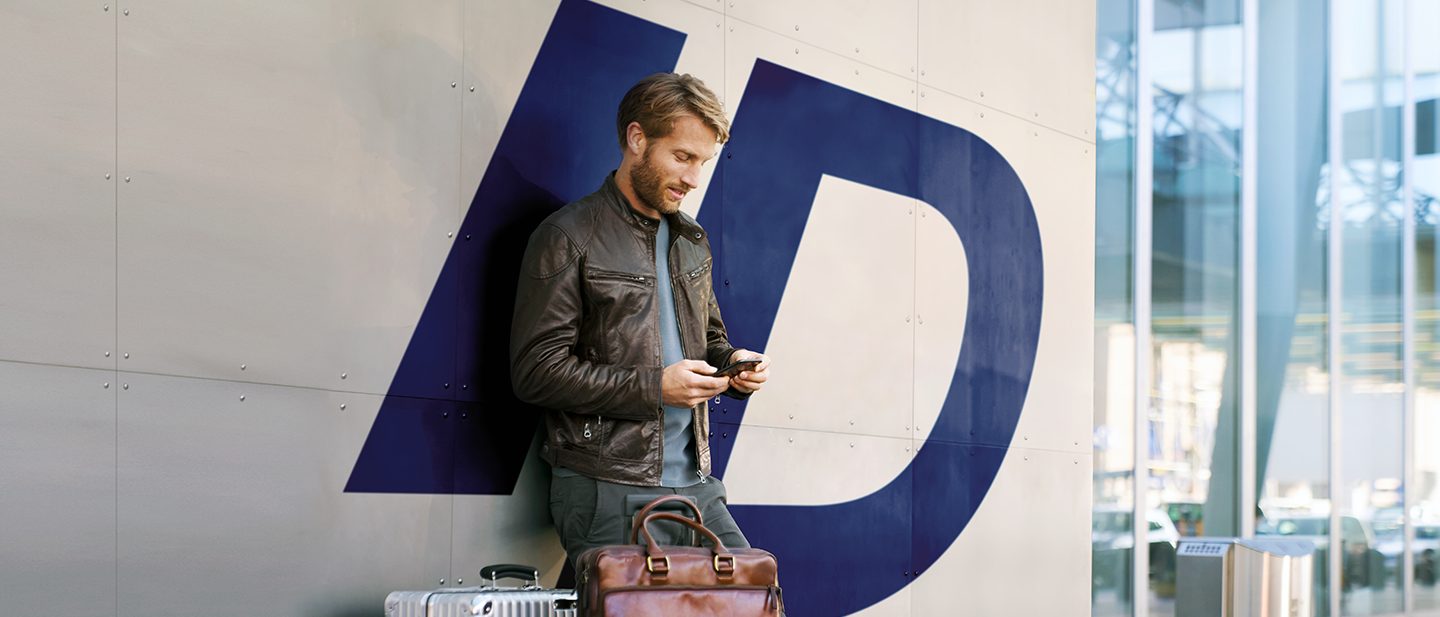Man in a leather jacket stands by luggage, checking his phone against a wall with blue lettering.