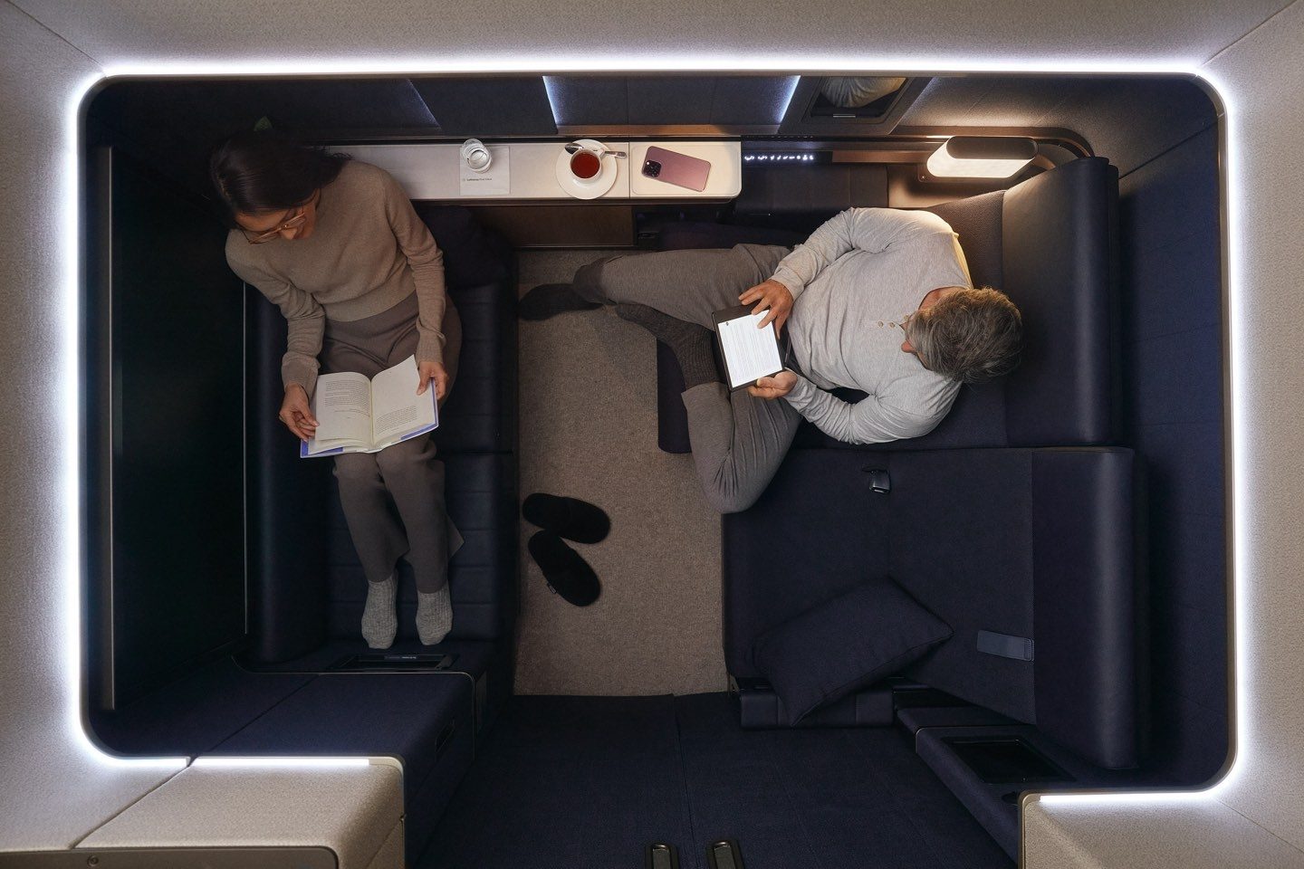 Two passengers relax in a premium airline suite, reading books & tablets.