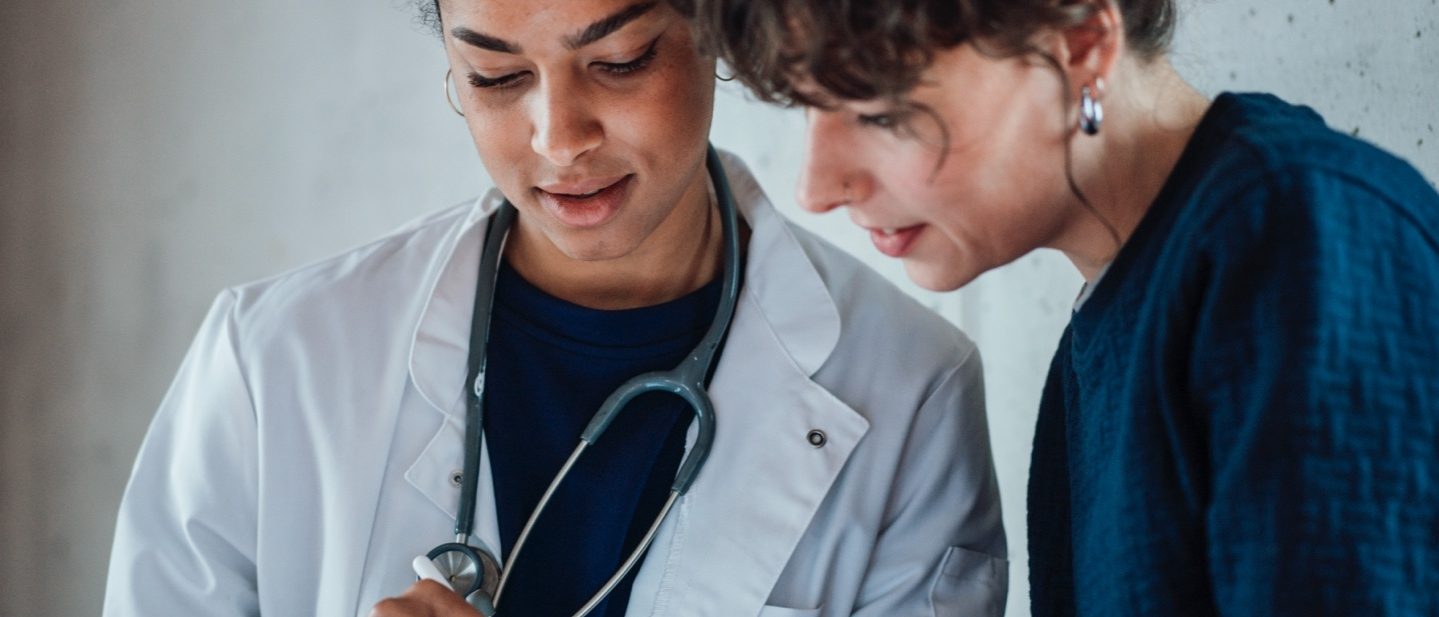 A woman doctor with a stethoscope and a female patient look at a tablet together.