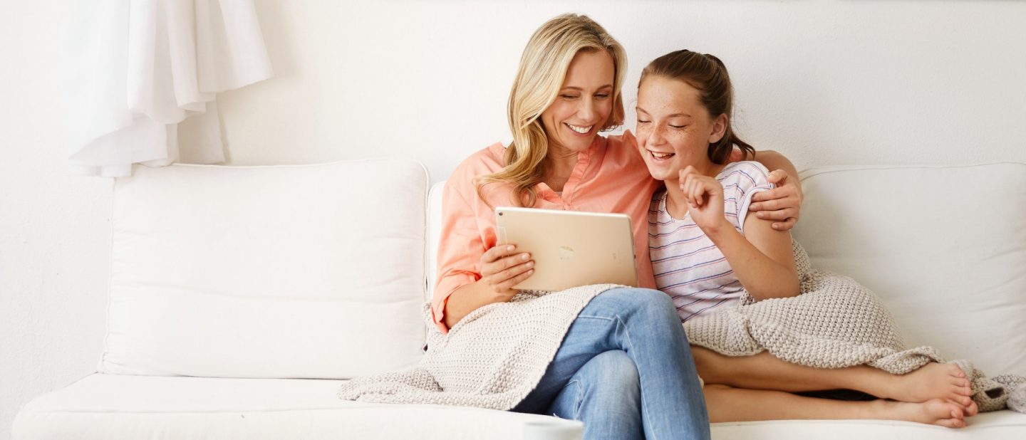 Mother and daughter smile on a sofa under a cozy blanket while looking at a tablet.