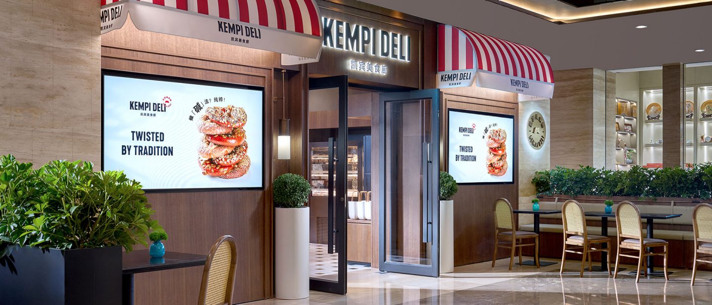 Entrance of the Kempi Deli restaurant with red-and-white striped awnings, illuminated display screens, and indoor seating.