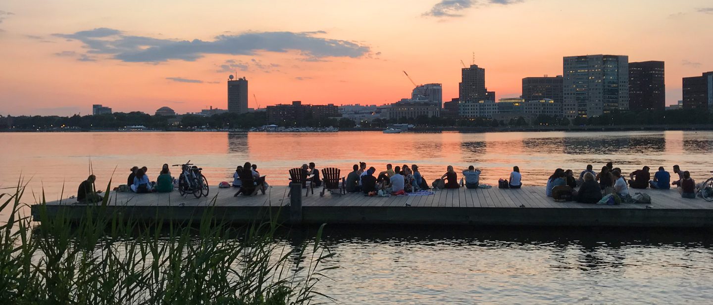 People sitting by the Charles River at sunset with the Boston skyline in the background