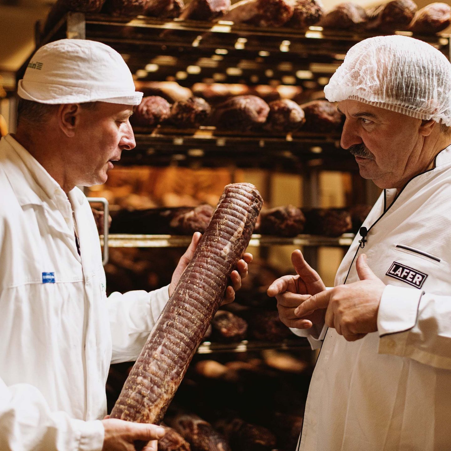 Two men in white coats and hairnets inspect cured meats in a storage room with racks of sausages.