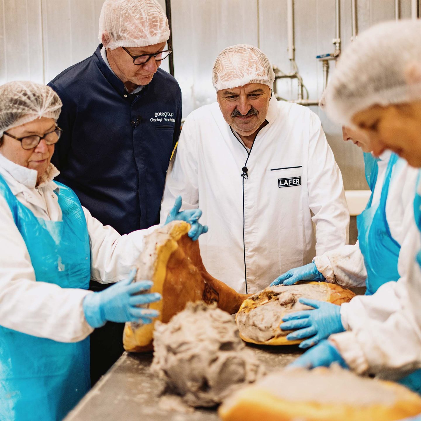 Four workers wearing hairnets and gloves inspect large cured hams in a meat processing facility.